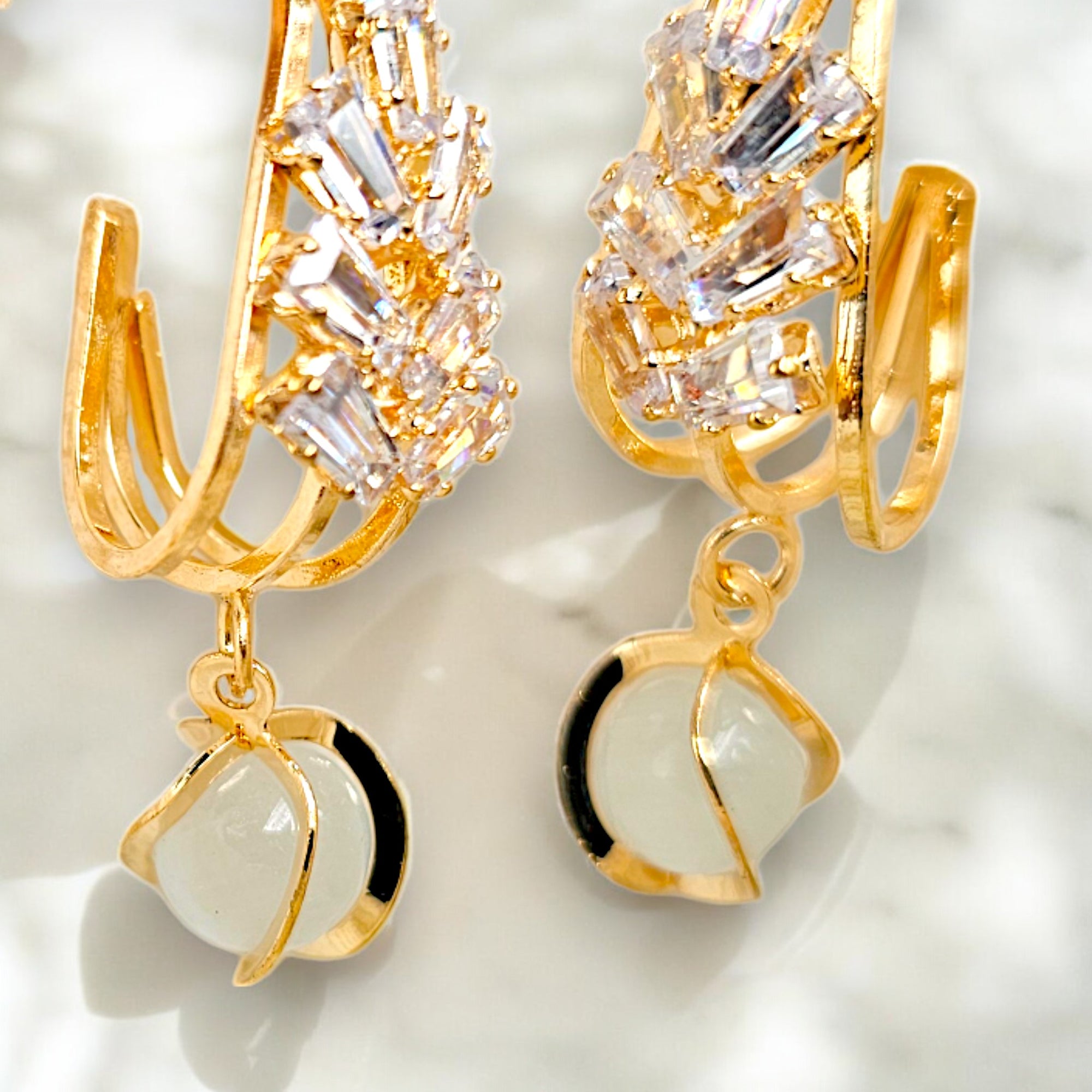 Elegant gold-plated dangle earrings featuring sparkling Zirconia crystals encased in a gold frame on a marble floor